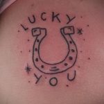 Фото простой татуировки подковы с надписью 'LUCKY YOU'. Минималистичный дизайн, контурный рисунок, татуировка на плече.