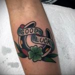Фото тату подковы и клевера с надписью 'Good Luck' в стиле олд скул на предплечье. Яркие цвета, четкие контуры, классический дизайн.