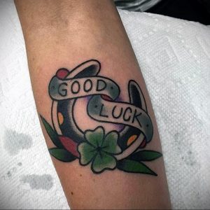 Фото тату подковы и клевера с надписью 'Good Luck' в стиле олд скул на предплечье. Яркие цвета, четкие контуры, классический дизайн.