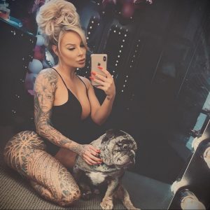 Фотография девушки с длинными светлыми волосами, татуировками на теле и мопсом, сидящим у нее на коленях.
