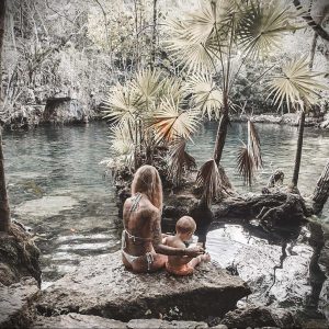 Фотография женщины с татуировками на теле и ее маленькой дочери, сидящих у воды. Женщина обнимает дочь.
