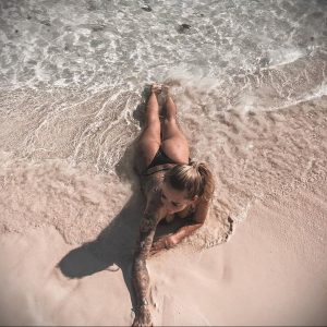 Фотография девушки с татуировками на руках, лежащей в воде у берега. Вид сверху демонстрирует татуировки на ее руках и плечах.