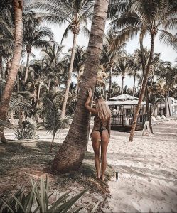 Фотография девушки с татуировками на теле, стоящей спиной к камере на тропическом пляже с пальмами.