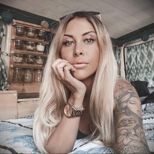 Фотография девушки с длинными светлыми волосами и татуировками на руках, смотрящей в камеру. Она опирается подбородком на руку.