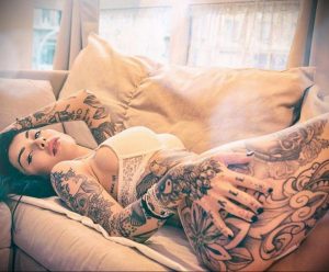 Фотография девушки с татуировками на руках и теле, лежащей на диване.