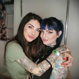 Фотография двух девушек с татуировками на руках, обнимающихся и улыбающихся.