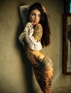 Фотография девушки с множеством татуировок на теле, одетой в белую рубашку, с соблазнительным взглядом.