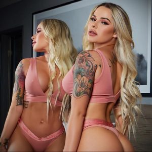 Фотография девушки с длинными светлыми волосами и татуировкой на плече и руке, позирующей в домашней обстановке.