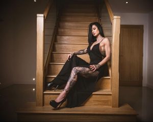 Фотография девушки в черном платье с детализированными татуировками на руках и ногах, сидящей на лестнице в роскошном интерьере.