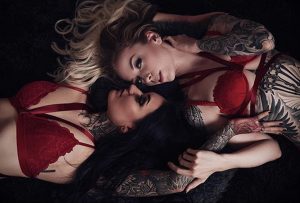Фотография двух девушек с татуировками, лежащих в постели в красном белье. Татуировки видны на руках, шее и теле.