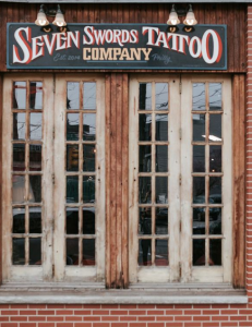 Фото фасада тату-салона "Seven Swords Tattoo Company" в Филадельфии. Видны деревянные двери и вывеска с названием салона.
