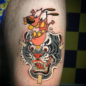 Фото яркой татуировки на руке, изображающей мультяшного пса Courage the Cowardly Dog, сражающегося с тигром. Использованы яркие цвета и четкие контуры.