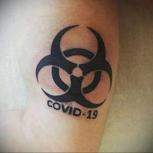 Фото тату с изображением знака биологической опасности и надписью 'COVID-19'. Черно-белая татуировка в минималистичном стиле.