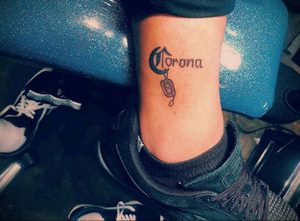 Фото тату с логотипом пива Corona, дополненным медицинской маской. Татуировка выполнена в черном цвете на лодыжке.