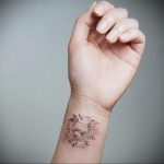 Фото тату мордочки лисы, окруженной цветами, на запястье. Рисунок выполнен в мягких пастельных тонах.