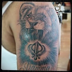 Фото тату льва на плече с изображением символа в центре. Рисунок выполнен в реалистичном стиле с использованием черных и серых оттенков.