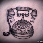 Фотография черно-белой татуировки старинного телефона с надписями 'Hey Boy' и 'Hey Girl' по бокам. Детализированный рисунок в стиле ретро.