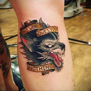 Фото тату волка на ноге с надписями 'This world owes you nothing'. Черно-серый рисунок в стиле олдскул с красными акцентами.