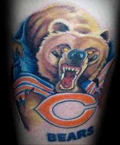 Фото тату медведя в спортивной форме с логотипом Chicago Bears на руке. Яркий рисунок татуировки с использованием синих, оранжевых и коричневых цветов.
