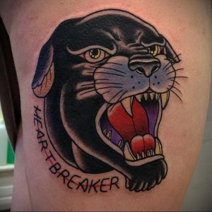 Фото татуировки пантеры с надписью "HEARTBREAKER" в стиле олд скул на бедре. Черный контур, яркие красные акценты, традиционный рисунок татуировки.