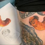 Фото тату: яркая и необычная татуировка куриной ножки с лицом на бедре. Рисунок татуировки выполнен в мультяшном стиле с использованием ярких оранжевых и желтых оттенков.