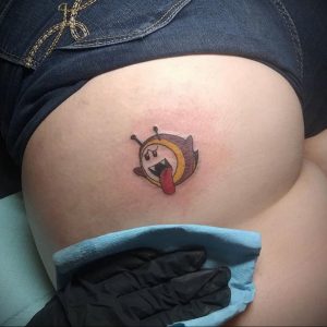 Фото тату на бедре с изображением мультяшного смайлика-пчелы с высунутым языком. Яркие цвета и игривый дизайн.