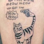 Фото тату на бедре с изображением кота, держащего пистолет, и надписью "You mess with the meow meow you get the peow peow", выполненное в черном цвете.