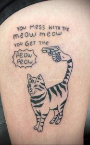 Фото тату на бедре с изображением кота, держащего пистолет, и надписью "You mess with the meow meow you get the peow peow", выполненное в черном цвете.
