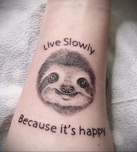 Фото тату на запястье с изображением милого ленивца и надписью "Live Slowly Because it's happy", выполненное в черно-белой графике.