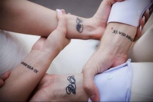Фото тату: На запястьях двух людей изображены римские цифры и небольшие символы, создающие парную татуировку.