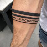 Фото татуировки на бицепсе мужчины, состоящей из римских цифр, заключенных между двумя черными полосами. Стильный и лаконичный дизайн.
