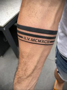 Фото татуировки на бицепсе мужчины, состоящей из римских цифр, заключенных между двумя черными полосами. Стильный и лаконичный дизайн.