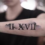 Фото тату: черные римские цифры 'II XVII' на предплечье, выполненные в минималистичном стиле с эффектом размытия по краям.