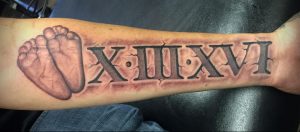 Фото татуировки на руке с римскими цифрами XIII XVI и отпечатком маленькой ножки. Черный цвет, реалистичный стиль, трогательный и значимый рисунок.