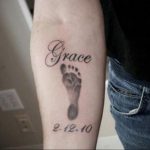 Фото татуировки на предплечье с отпечатком детской ноги и именем 'Grace'. Черно-серый цвет, реалистичный стиль, символизирует рождение ребенка.