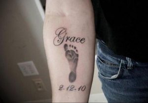 Фото татуировки на предплечье с отпечатком детской ноги и именем 'Grace'. Черно-серый цвет, реалистичный стиль, символизирует рождение ребенка.