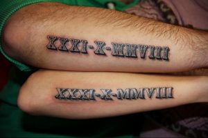 Фото парных татуировок на предплечьях с римскими цифрами 'XXXI-X-MMVIII'. Черный цвет, четкие линии, символизирует общую дату или событие.