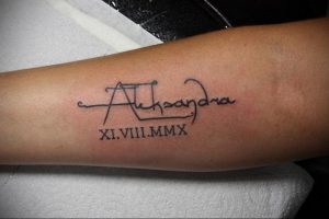 Фото татуировки на предплечье с именем 'Alexandra' написанным от руки и римской цифрой 'XI.VIII.MMX'. Черный цвет, каллиграфический стиль.