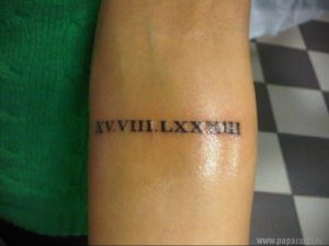 Фото татуировки на предплечье с римскими цифрами 'XV.VIII.LXXXIII'. Черный цвет, четкие линии, классический стиль.