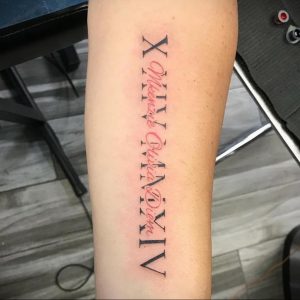 Фото тату: вертикальная татуировка римскими цифрами 'X Mkenzie Olivia Diem' и 'XIV' на предплечье. Черный цвет, каллиграфический шрифт.
