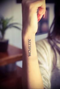 Фото тату: вертикальная татуировка римскими цифрами 'MCMLXIV' на запястье. Черный цвет, тонкие линии, минималистичный стиль.
