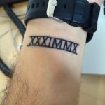Фото татуировки с римскими цифрами на запястье мужчины. Черный рисунок, простой и стильный дизайн, популярный выбор для памятных дат.