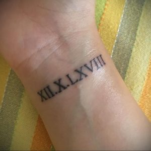 Фотография татуировки на запястье с римскими цифрами 'XII.X.LXVIII'. Черный цвет, тонкие линии, простой и элегантный дизайн.