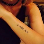 Фотография татуировки на предплечье с римскими цифрами 'I.XXIII.XCII'. Черный цвет, тонкие линии, изящный дизайн.