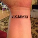 Фотография татуировки на запястье с римскими цифрами 'V.X.MMXI'. Черный цвет, тонкие линии, минималистичный дизайн.