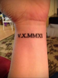 Фотография татуировки на запястье с римскими цифрами 'V.X.MMXI'. Черный цвет, тонкие линии, минималистичный дизайн.
