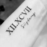 Черно-белое фото татуировки на предплечье с римскими цифрами 'XII.XCVII' и надписью 'Siztery'. Минималистичный дизайн, четкие линии.