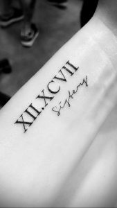 Черно-белое фото татуировки на предплечье с римскими цифрами 'XII.XCVII' и надписью 'Siztery'. Минималистичный дизайн, четкие линии.