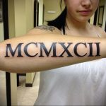 Фото крупной татуировки с римскими цифрами 'MCMXCII' на предплечье. Черный рисунок выглядит эффектно и привлекает внимание.