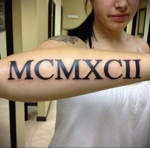 Фото крупной татуировки с римскими цифрами 'MCMXCII' на предплечье. Черный рисунок выглядит эффектно и привлекает внимание.